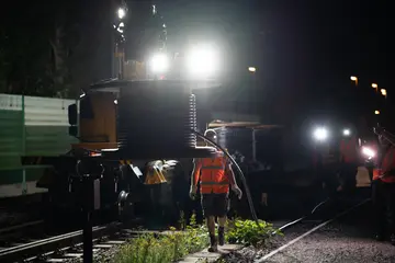 Das Symbolfoto zeigt eine Nachtbaustelle an einer Bahntrasse. Im Vordergrund ist eine Kabeltrommel zu sehen. Scheinwerfer erleuchten die Baustelle. Arbeiter der Deutschen Bahn/DB InfraGo besprechen sich.