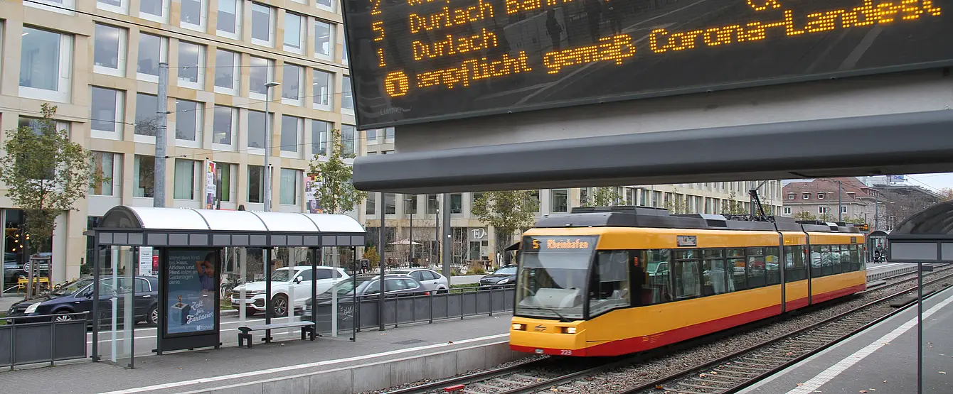 Eine Trambahn der Verkehrsbetriebe Karlsruhe fährt in die Haltestelle Tullastraße/Alter Schalchthof ein. Im Bildvordergrund ist ein Haltestellen-Anzeiger für die Fahrgastinformation zu sehen.