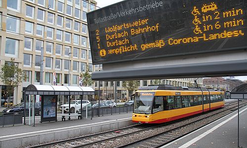 Eine digitale Fahrplan-Anzeige an einer Haltestelle. Im Hintergrund ist eine Trambahn der Linie 5 zu sehen.