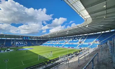 Das Foto zeigt aus der Perspektive der Südtribüne den Stadion-Innenraum im BBBank Wildpark mit Spielfeld und Zuschauertribünen. Weiße Wolken ziehen am blauen Himmel entlang.