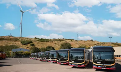 Zwölf neue E-Busse stehen in den VBK-typischen Farben gelb und rot vor einem Windrad.