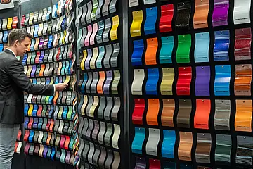 Ein Mann steht bei der Messe "Paint Expo" vor einer Wand mit lackierten Teilen der gesamten Farbpalette 