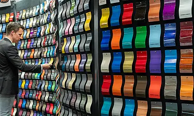 Ein Mann steht bei der Messe "Paint Expo" vor einer Wand mit lackierten Teilen der gesamten Farbpalette 