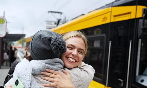 Zwei Frauen umarmen sich an einer Straßenbahnhaltestelle, an der eine Bahn hält.