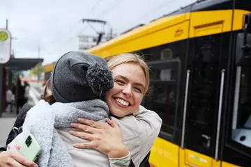 Zwei Frauen umarmen sich an einer Straßenbahnhaltestelle, an der eine Bahn hält.