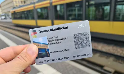 Eine Hand mit dem Deutschlandticket und einer Bahn im Hintergrund