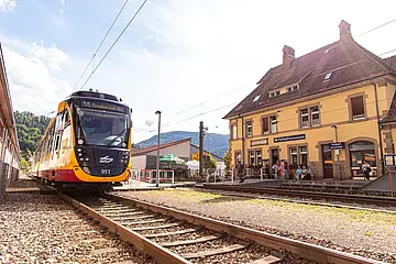 Eine Satdtbahn der AVG steht am Haltepunkt in Forbach. Im Hintergrund ist das historische Bahnhofsgebäude zu sehen.