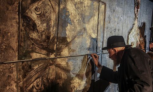 Markus Lüpertz steht vor einem Wandrelief des Gesamtkunstwerkes "Genesis" und arbeitet mit einem Werkzeug in der Hand einige Linien am Relief nach.