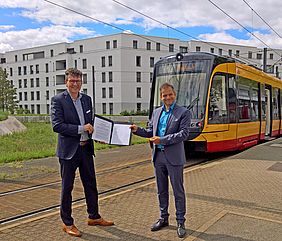 Geschäftsführer Stefan Storz und KVV-Geschäftsführer Dr. Alexander Pischon stehen vor einer Bahn und halten gemeinsam einen Vertrag in den Händen
