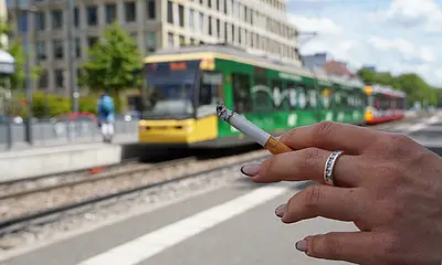 Eine Hand hält eine Zigarette an einer Bahnhaltestelle. Im Hintergrund fährt eine Straßenbahn vorbei