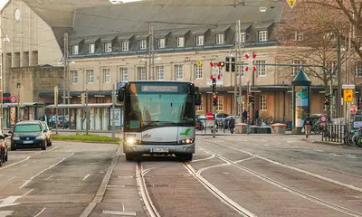 Ein Linienbus fährt auf einer Straße, in der auch Bahngleise liegen. Im Hintergrund ist der Karlsruher Hauptbahnhof zu sehen.