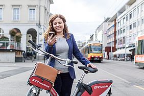 Lächelnde junge Frau hält ein Smartphone in der Hand und steht hinter einem rotem KVV.nextbike. Im Hintergrund ist eine Stadtbahn zu sehen.