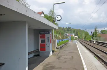 Der Bahnsteig am Haltepunkt Bilfingen: In der Wartehalle in der linken Bildhälfte ist ein Fahrkartenautomat zu sehen. Am rechten Bildrand verlaufen die Gleise der Bahnstrecke. Der Himmel ist bewölkt.