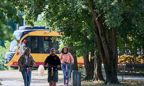 Drei junge Menschen fahren auf Rollern von Voi durch Karlsruhe. Im Hintergrund ist eine Stadtbahn zu sehen.