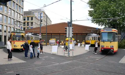 Historische Straßenbahnen stehen vor einer Wagenhalle auf dem betriebsglände der Verkehrsbetriebe Karlsruhe.. Menschen schauen sich die Fahrzeuge an.