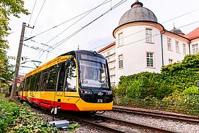 Eine gelb-rote Straßenbahn fährt an dem Schloss in Ettlingen vorbei. Der Himmel ist leicht bewölkt.