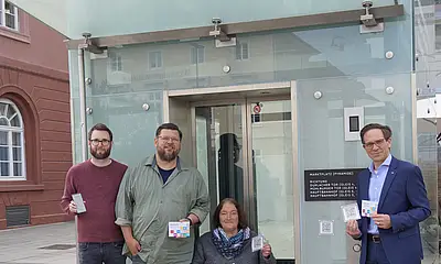 Vor dem Stadtbahn-Tunnel am Marktplatz sind von rechts nach links folgende Personen zu sehen: Christian Höglmeier (technischer Geschäftsführer der VBK), Ulrike Wernert (kommunale Behindertenbeauftragte der Stadt Karlsruhe), Artur Bundnik (Projektstelle Web-App „Karlsruhe barrierefrei“) und Marcus Albert (System-Administrator „Karlsruhe barrierefrei“). Sie halten die Aufkleber mit den QUR-Codes zu "Karlsruhe barrierefrei" in der Hand. Vor ihnen sitzt ein Hund. 