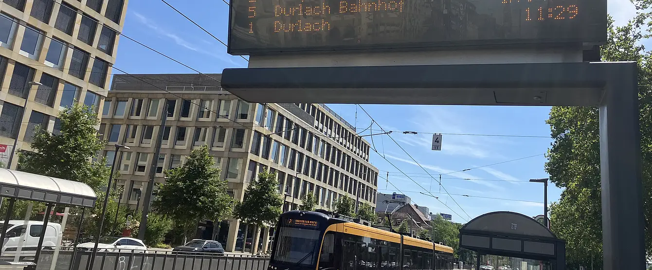 Eine Trambahn der Verkehrsbetriebe Karlsruhe fährt in die Haltestelle Tullastraße/Alter Schalchthof ein. Im Bildvordergrund ist ein Haltestellen-Anzeiger für die Fahrgastinformation zu sehen.