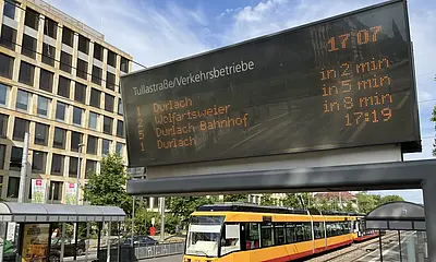 Eine Trambahn der Verkerhsbetriebe Karlsruhe fährt in eine Haltestelle ein. Im Vordergrund ist ein Haltestellen-Anzeiger zur Fahrgastinformation zu sehen.
