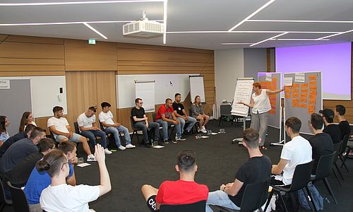 Junge Menschen sitzen in einem stuhkreis in einem Seminarraum und diskutieren mit einer Referentin über das Thema Nahchhaltigkeit.
