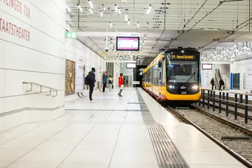 Das Foto zeigt die Stadtbahntunnel-Haltestelle am Ettlinger Tor/Staatstheater in Karlsruhe. Eine Bahn fährt in die Haltestelle ein. Am Bahnsteig stehen Menschen
