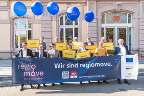 Die Projektpartnervon regiomove halten ein blaues Banner mit der Aufschrift "Wir sind regiomove". Dahinter sind blaue regiomove-Luftballons und das Bahnhofsgebäude in Baden-Baden zu sehen.