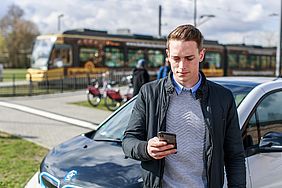 Ein Mann hält ein Smartphone in der Hand. Im Hintergrund sind ein Auto, Leihfahrräder und eine Straßenbahn zu sehen.