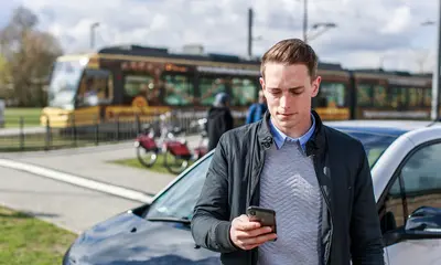 Ein junger Steht mit einem Smartphone in der Hand vor einem Carsharing-Auto. Im Hintergund sind Leihfahrräder und eine Straßenbahn zu sehen.