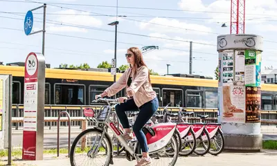 Brunette Frau fährt auf einem Fahrrad der Firma nextbike. Im Hintergrund sind weitere Fahrräder und eine gelbe Straßenbahn zu sehen. Der Himmel ist blau und leicht bewölkt.