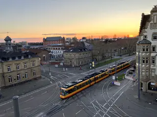 Das Foto zeigt aus der Vogelperspektive das Gleisviereck Tullastraße/Durlacher Allee mit einer vorbeifahrenden Straßenbahn. Im Hintergrund ist das Schloss Gottesaue zu sehen. Die Sonne geht gerade unter.