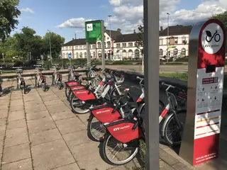 Räder des Bike-Sharing-Angebots kvv.netbike stehen an einer Ausleihstation auf dem Platz an der Reithalle in Rastatt. Im Hintergrund ist das Bahnhofsgebäude von Rastatt zu sehen