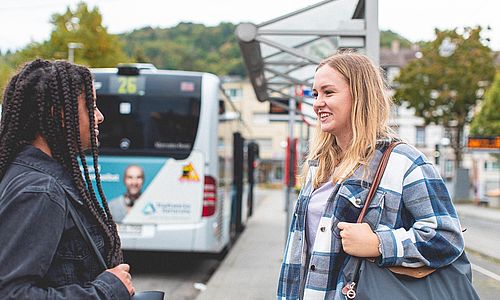 Zwei Schülerinnen stehen an einer Haltestelle vor einem Bus.