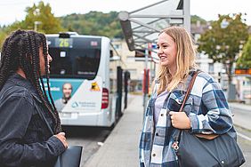 Zwei Schülerinnen stehen an einer Haltestelle vor einem Bus.