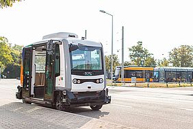 Am Straßenrand steht ein autonom fahrender, scharz-weißer Mini-Bus mit geöffneter Tür. Im Hintergrund sind die Haltestelle Weiherfeld-Dammerstock und eine gelbe Stadtbahn zu sehen.