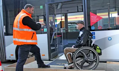 Ein Mitarbeiter der Verkehrsbetriebe Karlsruhe gibt einem Fahrgast, der in einem Rollstuhl sitzt, Tipps für den sicheren Ein- und Ausstieg bei einem Linienbus der VBK.