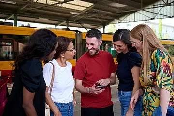 Junge Menschen stehen an einer Bahnhaltestelle und schauen gemeinsam auf ein Smartphone.