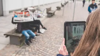Eine Schülerin hält ein Tablet in der Hand. Im Bildhintergrund sitzen zwei Schüler mit einer Zeitung auf einer Bank.