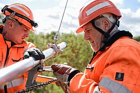 Zwei Männer in orangefarbener Kleidung reparieren eine Oberleitung.