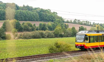 Eine AVG-Stadtbahn fährt auf der Kraichgaubahn. Im Hintergrund sind bewaldete Hügel zu sehen