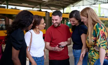 Fünf Menschen stehen an einer Haltestelle. Ein Mann hält ein Smarthone in der Hand. Im Hintergrund ist eine Stadtbahn zu sehen.